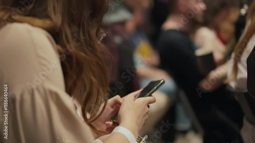 Young adult woman with long hair wearing glasses writes notes for speech into her phone. Attendees use smartphones to document a speech on cutting-edge technologies and innovations in packed venue