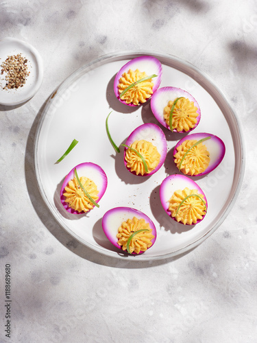 Delicious deviled eggs are beautifully arranged on a round white plate, featuring vibrant purple edges. Each egg is garnished with fresh herbs and served alongside a small dish of seasoning photo