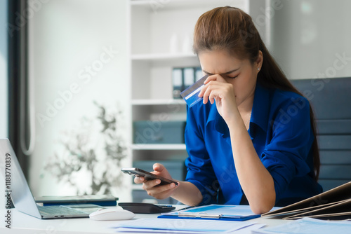 An Asian businesswoman in a blue formal shirt is working at her desk, analyzing financial charts on a laptop and mobile phone, focusing on credit management and financial strategies. photo