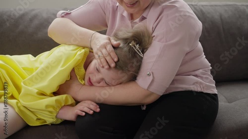 Young girl with Down syndrome rests on mother's lap while being gently patted on head. Scene captures moment of tenderness and care, highlighting emotional support. Concept: well-being, family love
