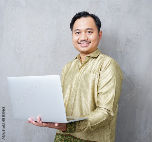 Adult Asian man wearing batik smiling at camera while using his laptop - Indonesia men photo