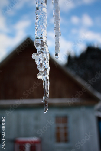 Transparent icicle close-up. There is a blurry image of an old village house in the background.