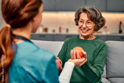 Smiling senior woman holding a spiked ball in her hand for rehabilitation and recovery of arm muscles after illness. Nurse supports and helps. Physical therapy and recovery.