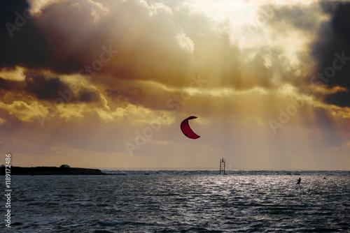 
A kitesurfer riding the sea using the power of the wind, with a ship and a sky with clouds behind him.