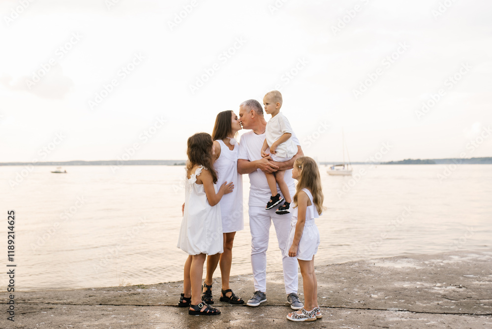 Happy family is standing on beach with boat in the background. Father is holding baby and other children are standing around him and kiss mother Happy family is standing on beach with boat in the background. Father is holding baby and other children are standing around him and kiss mother