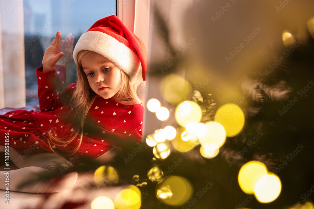 Little girl with Santa hat near window in room decorated for Christmas Little girl with Santa hat near window in room decorated for Christmas