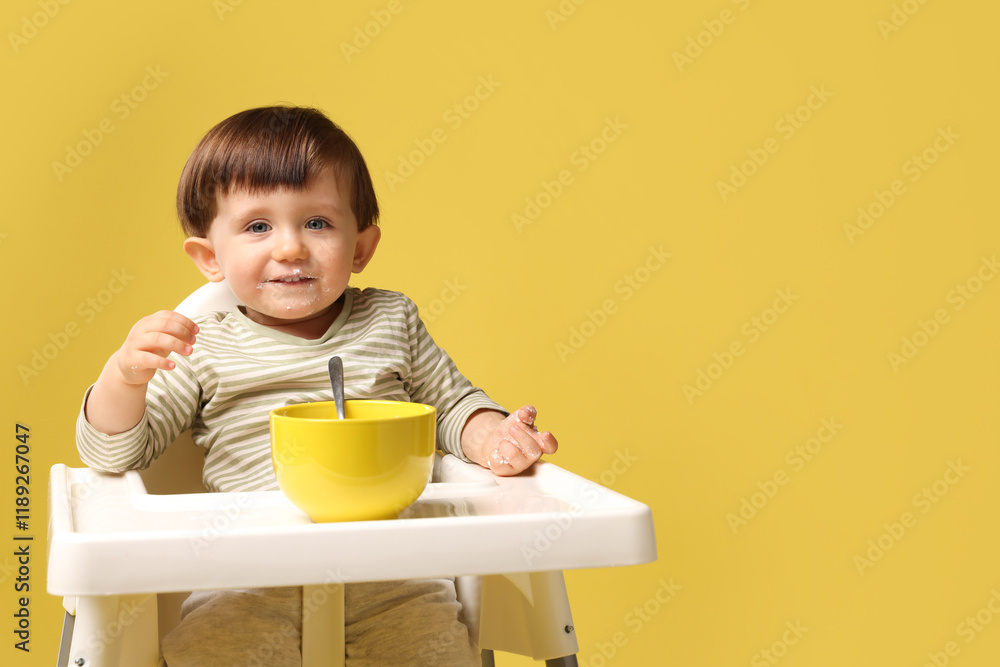Cute little kid eating healthy baby food from bowl in high chair on yellow background, space for text Cute little kid eating healthy baby food from bowl in high chair on yellow background, space for text