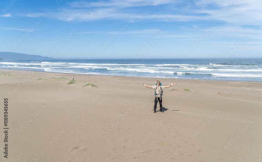 A young backpacker with open arms, standing on a beach, posing for a photo while admiring the stunning coastal landscape.