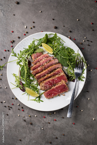 Grilled tuna steak with sesame seeds, cut into pieces with arugula and lime on a white plate on a gray background, top view.