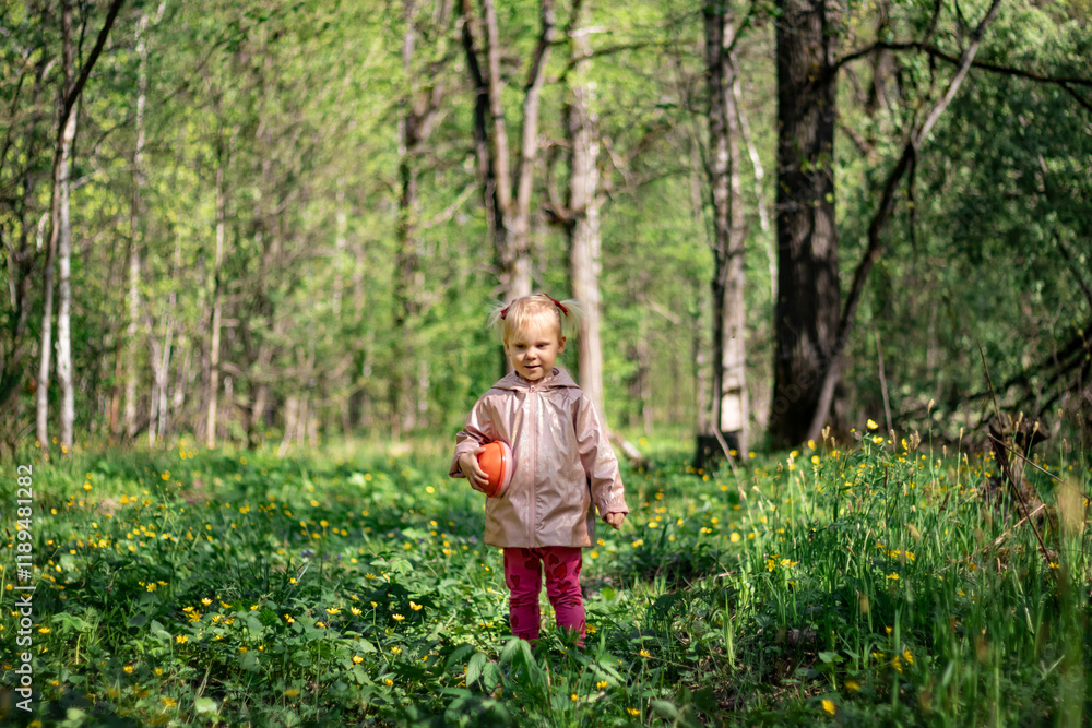 A little girl of 2-3 years old stays in the forest and enjoys nature. Background of a green forest. The girl holds tightly in her hand an orange jar in which she collects the flowers she likes. She is
