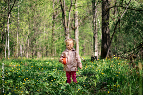 Obraz na płótnie A little girl of 2-3 years old stays in the forest and enjoys nature. Background of a green forest. The girl holds tightly in her hand an orange jar in which she collects the flowers she likes. She is w salonie