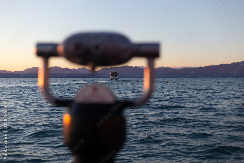A boat approaches under a warm sunset, framed artistically by a blurred telescope and serene mountain scenery.