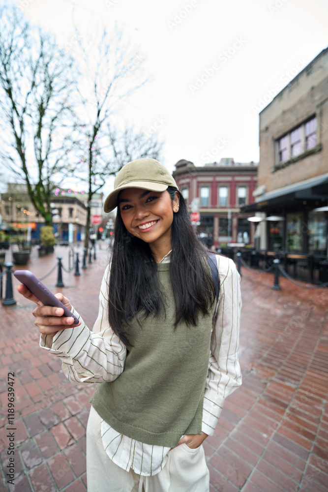 Fototapeta premium Smiling cool teen girl holding mobile cell phone having fun outdoors with cellphone in hands. Happy Asian teenager using smartphone chatting online looking at camera in the city. Vertical portrait.