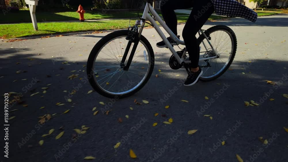 A young woman riding bicycle at the street in suburbs neighborhood on a beautiful bright sunny autumn day. Many yellow leaves are on the road. A young woman riding bicycle at the street in suburbs neighborhood on a beautiful bright sunny autumn day. Many yellow leaves are on the road.
