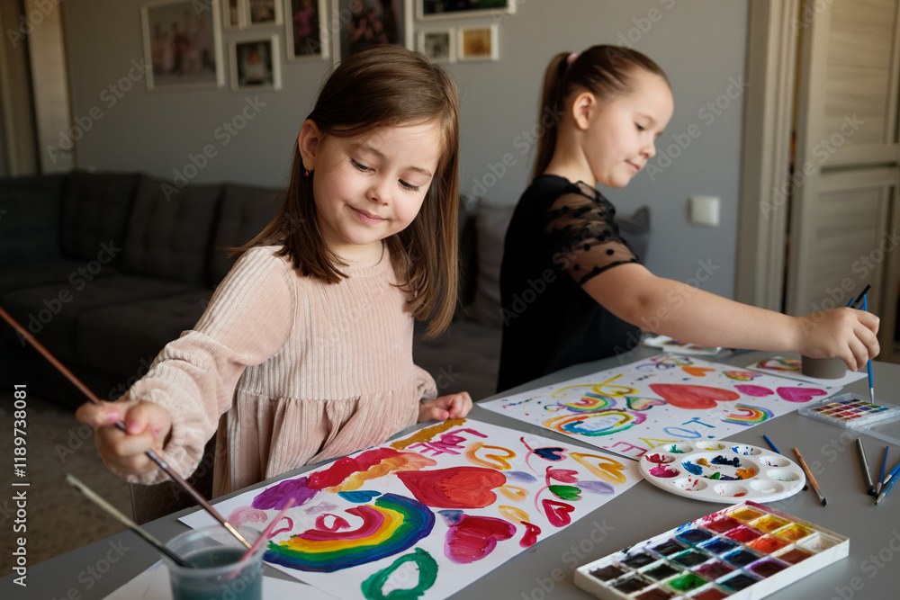 Fototapeta premium Two children are happily painting with watercolors, expressing creativity in a warm, homey environment. Art supplies are neatly arranged on the table