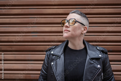 Short-haired woman wearing sunglasses and leather jacket posing near brown background photo