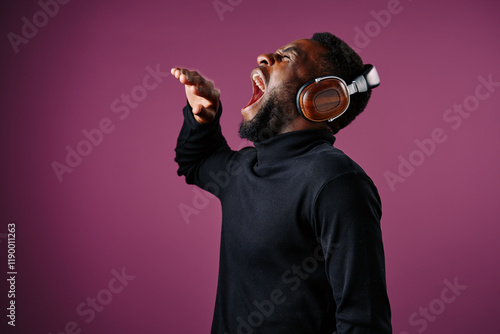 Young African man in black turtleneck energetically singing while wearing headphones against a purple background, expressing passion for music and creativity photo