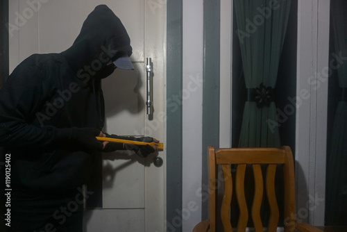 Illustration: A thief wearing all black and a hood trying to get into a house by prying open the door using a crowbar at night. photo