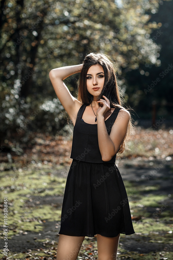 A girl with long dark hair standing against the background of a forest. She is wearing a sleeveless black dress A girl with long dark hair standing against the background of a forest. She is wearing a sleeveless black dress