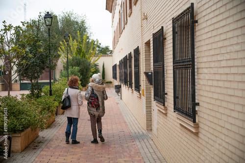 Wallpaper Mural Two senior women walking through a peaceful pedestrian street surrounded by greenery and traditional brick architecture Torontodigital.ca