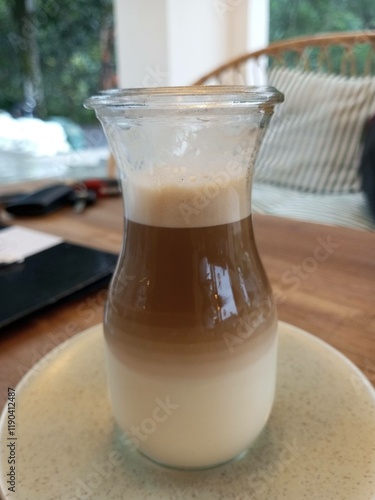 A close-up of a beautifully layered latte served in a transparent glass cup on a ceramic saucer, showcasing the separation of milk and coffee with foam on top.