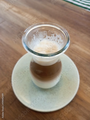 A close-up of a beautifully layered latte served in a transparent glass cup on a ceramic saucer, showcasing the separation of milk and coffee with foam on top.