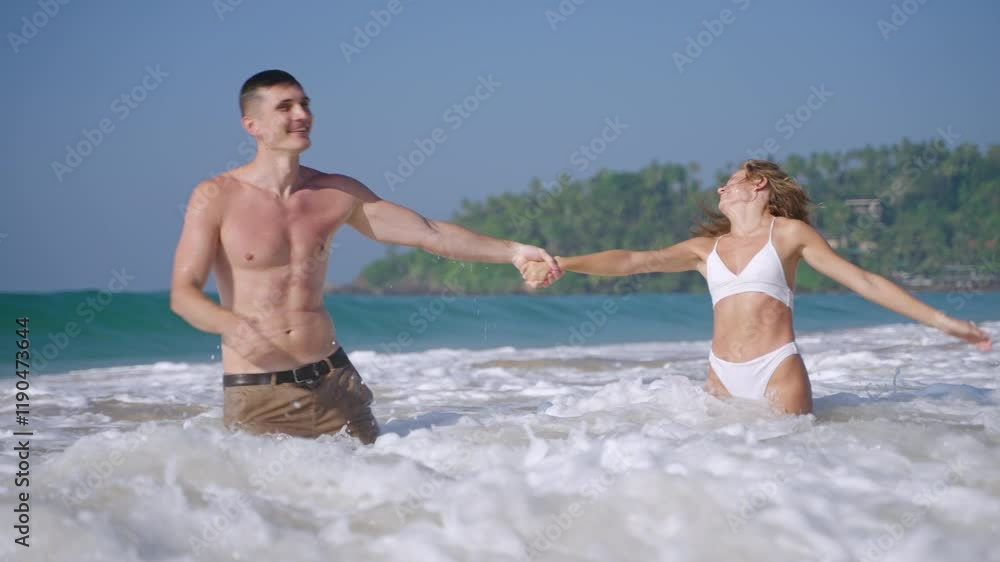 Young fit couple holding hands, having fun among the waves in the ocean, water splashes and foam. Smiling boyfriend and girlfriend playing in sea. Sporty man, woman have a romantic trip in summer. Young fit couple holding hands, having fun among the waves in the ocean, water splashes and foam. Smiling boyfriend and girlfriend playing in sea. Sporty man, woman have a romantic trip in summer.