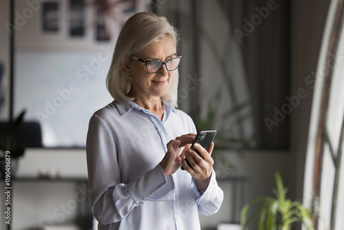 Serious middle-aged businesslady using on-line app on smartphone, enjoying modern technology, wireless internet, remote communication, typing text messages to client, smiling, look at cellphone screen photo