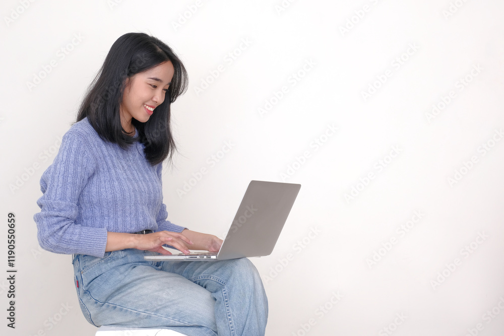 Side view: A young woman sitting on a wooden bench, working on a laptop on her lap with a smiling, happy expression Side view: A young woman sitting on a wooden bench, working on a laptop on her lap with a smiling, happy expression