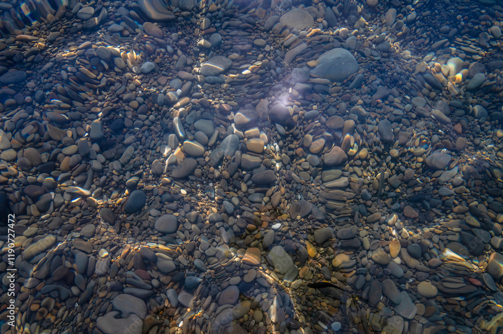 Underwater pebble backgrounds. Colorful stones sea bottom in the sun rays. View from above through clear water. Seascape wallpaper texture. Defocus.
