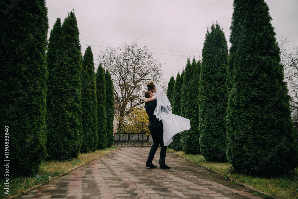 A bride and groom are standing in a garden with a white veil in the air. The bride is holding the groom's arm while they both look up. Scene is romantic and happy A bride and groom are standing in a garden with a white veil in the air. The bride is holding the groom's arm while they both look up. Scene is romantic and happy