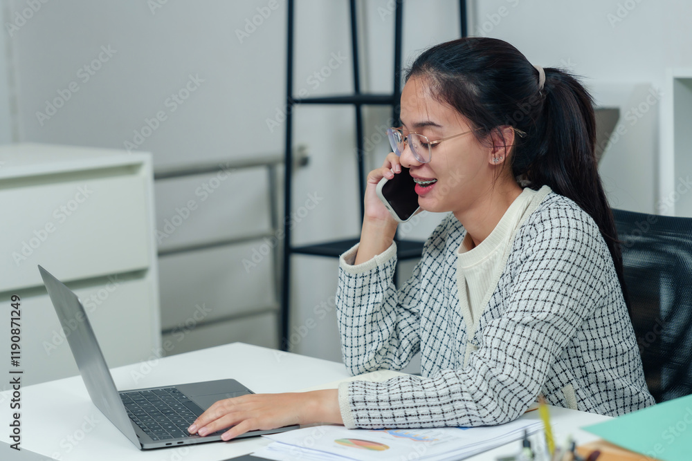 Young Asian office worker talking on phone while typing on laptop, smiling during conversation in organized workspace, focusing on multitasking and professional communication Young Asian office worker talking on phone while typing on laptop, smiling during conversation in organized workspace, focusing on multitasking and professional communication