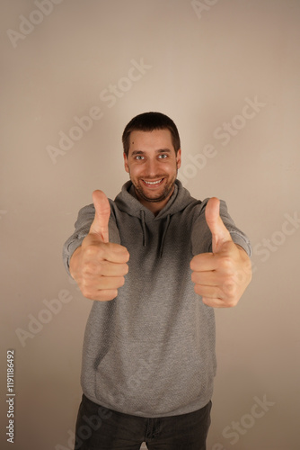 He s a brunette of European descent, wearing a grey hoodie. His casual style makes him look relaxed and approachable. A cheerful man shows a thumbs-up with both hands, radiating positive energy. photo