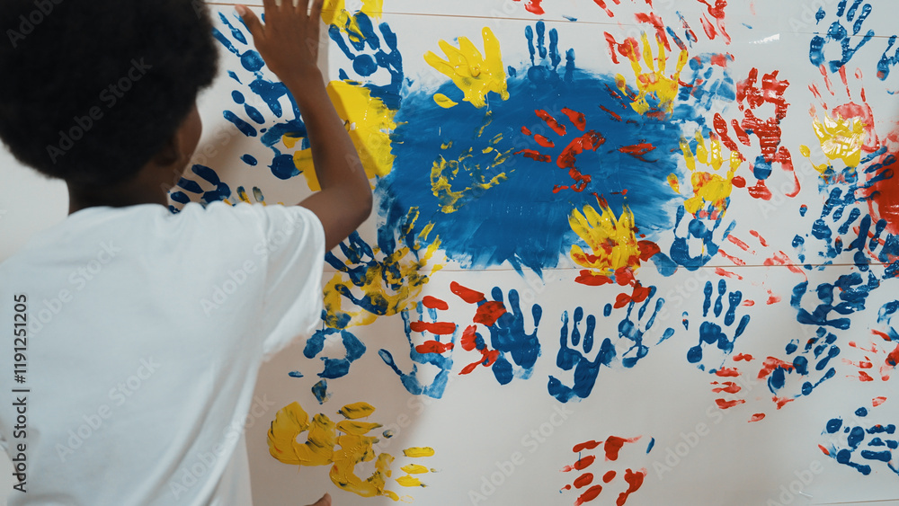 Obraz premium Back view of happy african boy painted the stained wall with colorful hand while wearing casual white shirt in art lesson.Smart student use hand print to make creative artwork. Education. Edification.