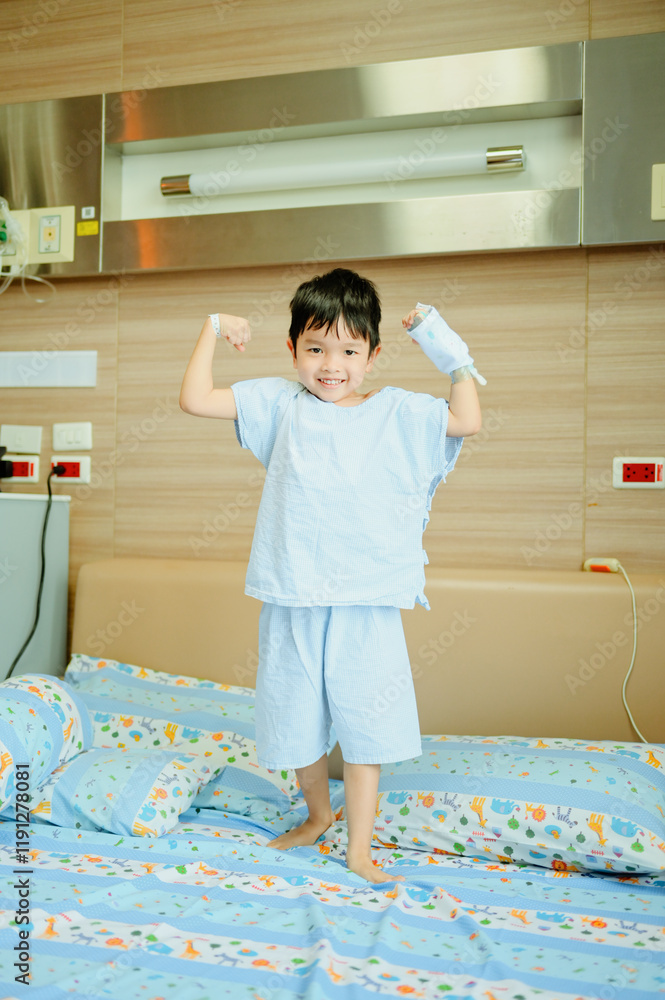 Young boy wearing light blue hospital pajamas, standing on a bed with a cheerful expression