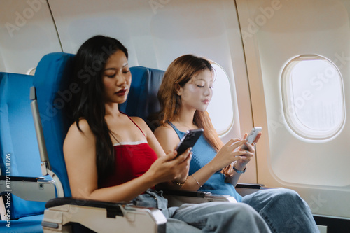 Two young women are sitting in airplane seats, using their smartphones during a flight, possibly browsing the internet, checking social media, or playing games