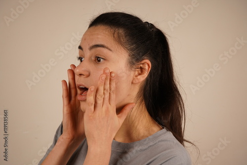 A young woman places her hands to her cheeks, her eyes wide open in genuine shock. The expression vividly captures a moment of astonishment and surprise. Her pose emphasizes the unexpected reaction.