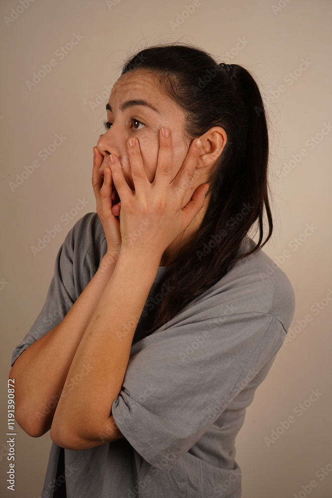 The interesting facial expression conveys a moment of surprise. The focus is on her emotions. A young woman holds her hands over her face, her eyes wide open and her mouth open in genuine surprise. The interesting facial expression conveys a moment of surprise. The focus is on her emotions. A young woman holds her hands over her face, her eyes wide open and her mouth open in genuine surprise.