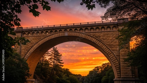 A medium closeup of the arch of an arch bridge during sunset capturing the warm hues illuminating the stonework while silhouetted trees frame the scene. photo