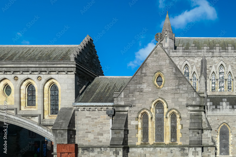 Obraz premium Section of a grey stone building of Christ Church Cathedral in Dublin, Ireland. Blue sky with clouds. Pointed arches frame tall windows with stained glass. Steeply pitched slate roof leads to spire