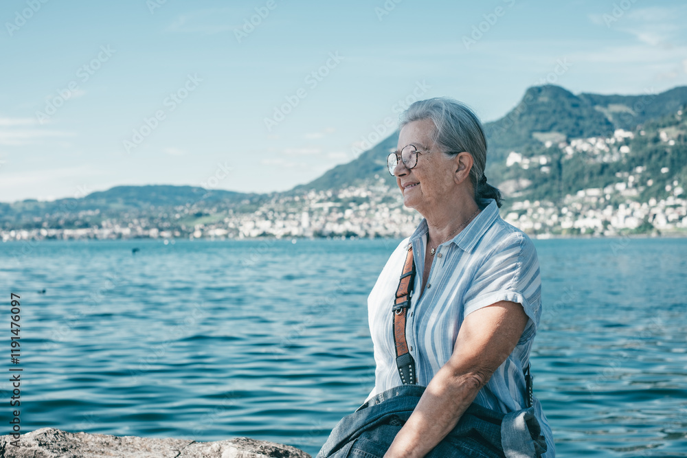 Fototapeta premium Senior white haired woman sitting along lake admiring the landscape - relaxed senior grandmother enjoying peaceful moments in nature, Lake Geneva and mountains in background