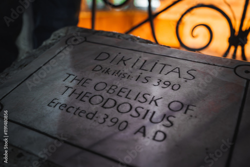 Close up of a marble plaque inscribed with details of the Obelisk of Theodosius, framed by ornate wrought iron railing and warm orange lighting. photo