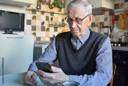 Attractive elderly man, 85 years old, looking at smartphone while sitting in kitchen at home