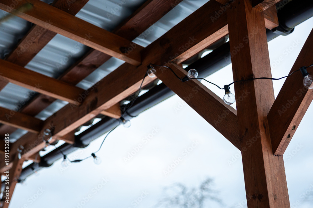 Obraz premium Outdoor wooden terrace structure with a corrugated metal roof, featuring string lights with round bulbs and a pale sky with tree branches in the background.