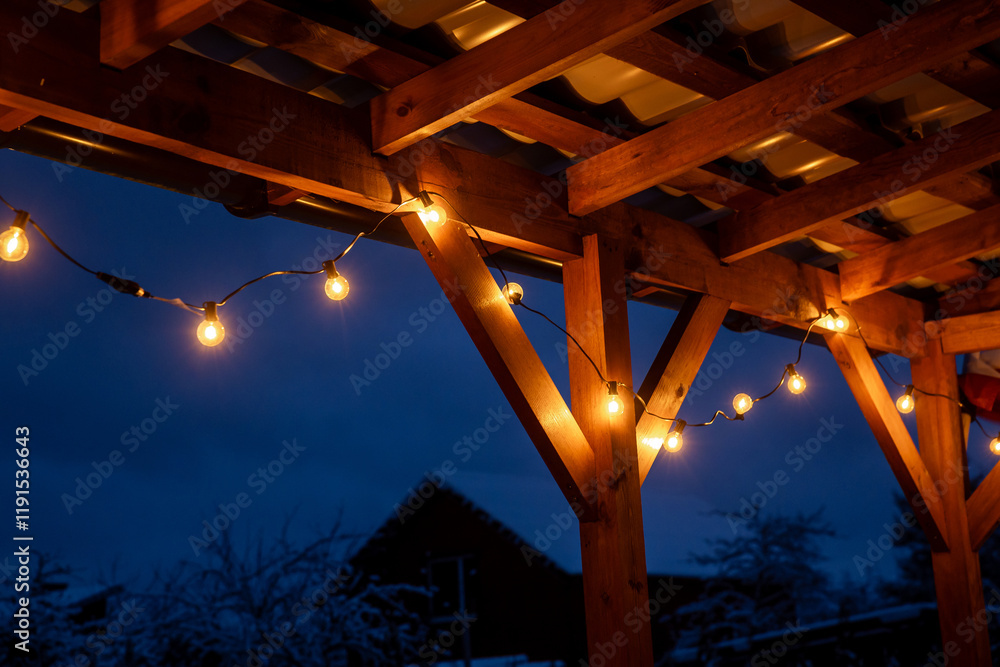 Obraz premium String lights with yellow bulbs hang on a wooden terrace with a corrugated roof. Snow covers the ground and trees under a deep blue evening sky.