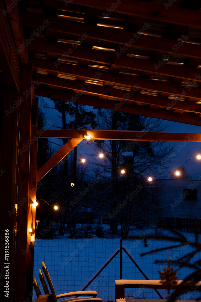 Obraz premium A terrace with warm LED string lights on a wooden pergola, wooden furniture, and a snow covered yard with trees and a building at dusk.