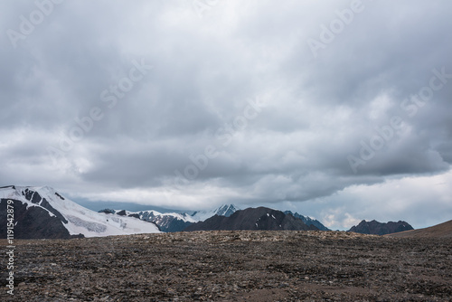 Dramatic misty top view from stone pass to big glacier tongue among sharp rocks and large snow-capped pinnacle in rainy low clouds. Dark atmospheric mountain silhouettes in rain in gray cloudy sky. photo