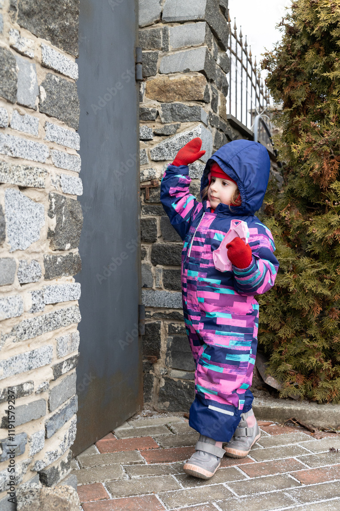 A little girl stands near a stone wall and curiously explores the surroundings. The child is dressed in a warm winter jumpsuit. The child holds a toy in her hand. A little girl stands near a stone wall and curiously explores the surroundings. The child is dressed in a warm winter jumpsuit. The child holds a toy in her hand.