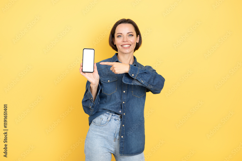 A young woman with a brunette hairstyle is standing against a vibrant yellow background. She is casually dressed and smiling while pointing at her smartphone, highlighting its screen. A young woman with a brunette hairstyle is standing against a vibrant yellow background. She is casually dressed and smiling while pointing at her smartphone, highlighting its screen.