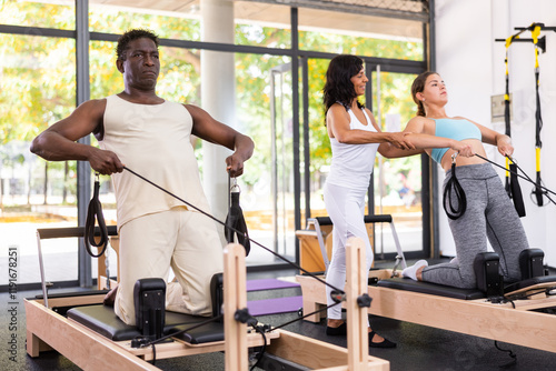 Hispanic woman pilates trainer correcting moves of young cacuasian woman and african-american man who doing exercises on reformers. photo